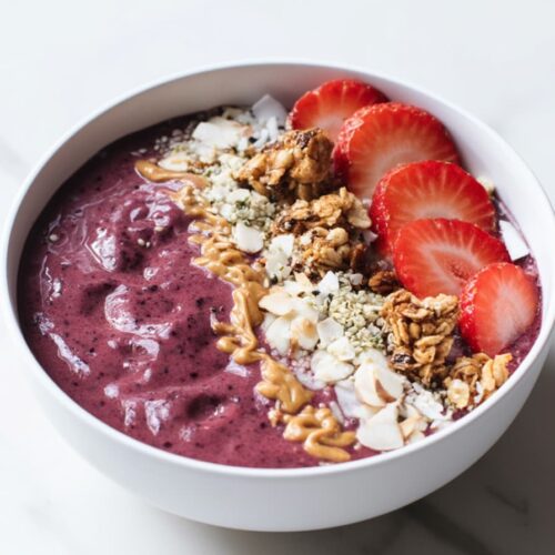 Thick berry protein smoothie bowl made with blueberries, strawberries, and raspberries, topped with sliced strawberries, granola, coconut flakes, almonds, hemp seeds, and peanut butter drizzle in a white bowl