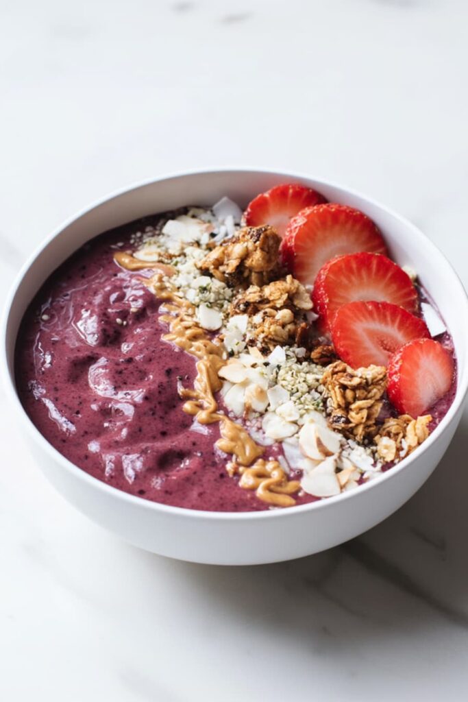 Thick berry protein smoothie bowl made with blueberries, strawberries, and raspberries, topped with sliced strawberries, granola, coconut flakes, almonds, hemp seeds, and peanut butter drizzle in a white bowl