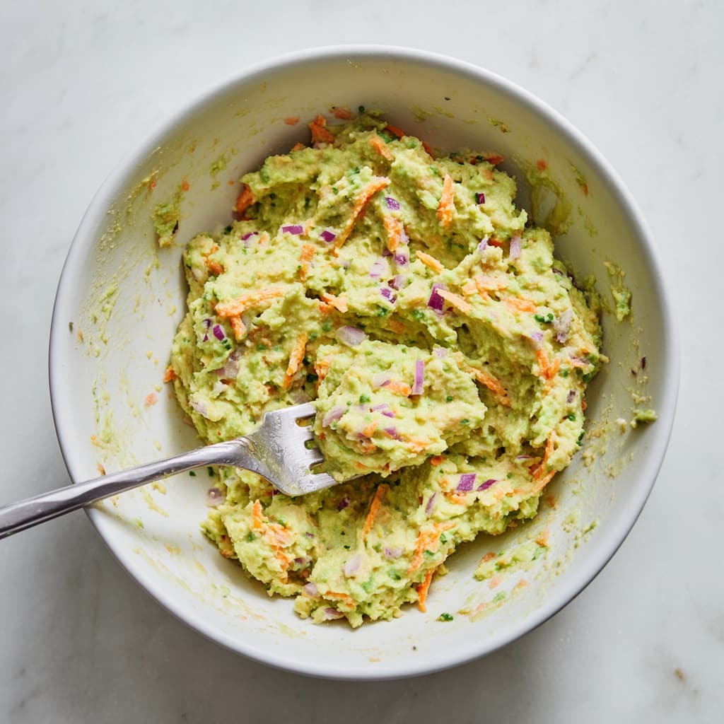 Chopped red onion and shredded carrots added to creamy chickpea avocado mixture in a bowl.