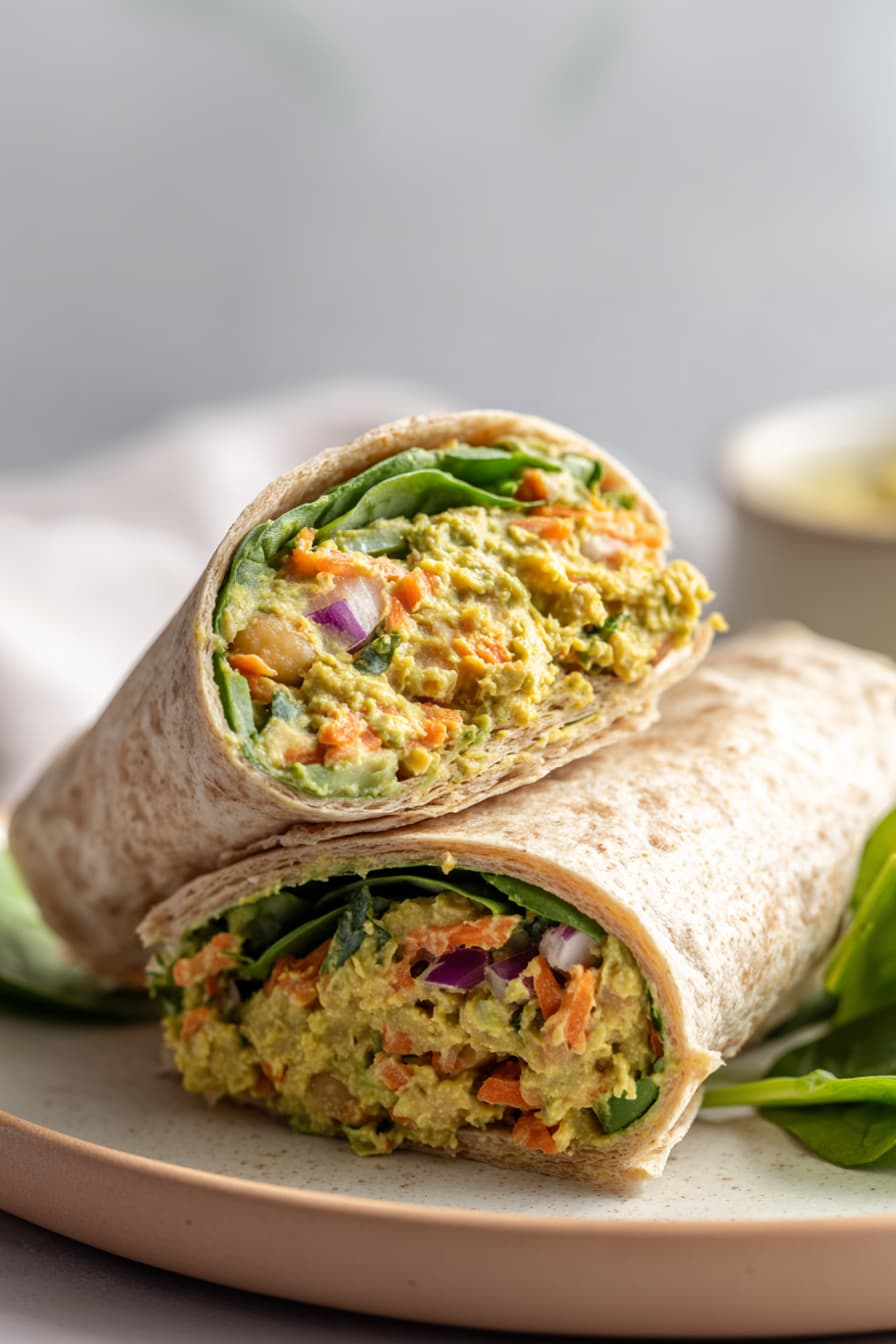 Chickpea avocado salad wrap cut in half showing creamy chickpea filling, carrots, red onion, and spinach inside whole grain tortilla.