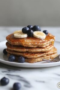 Stack of fluffy high-protein cottage cheese pancakes topped with fresh fruit, Greek yogurt, and a drizzle of maple syrup on a white plate.