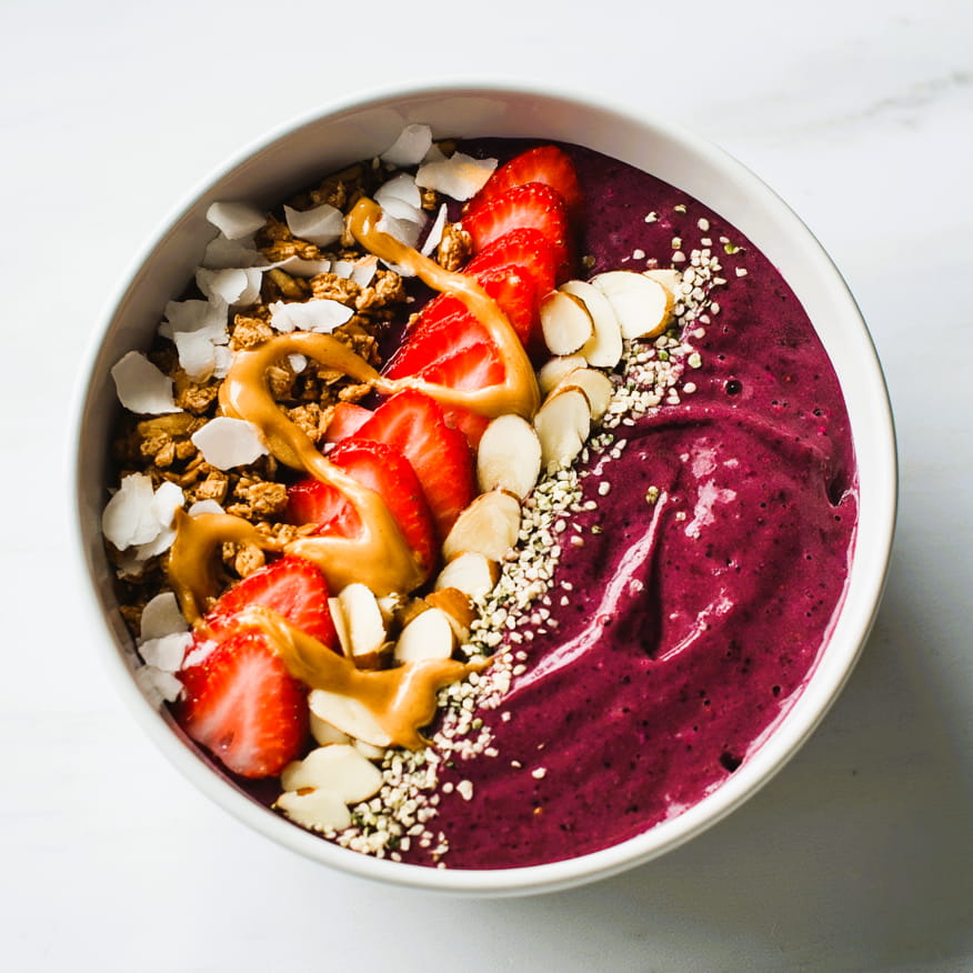 finished berry protein smoothie bowl with neatly arranged toppings, spoon resting inside bowl, thick texture visible, vibrant purple base with colorful toppings, white ceramic bowl on marble surface, soft natural daylight, clean fitness food aesthetic, realistic food photography, high detail, same bowl and setup