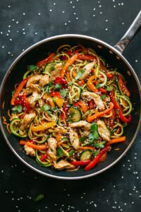 Healthy chicken stir-fry with zucchini noodles, red and yellow bell peppers, and carrots in a sesame garlic sauce, garnished with cilantro and sesame seeds—low carb, high protein, and perfect for clean eating.