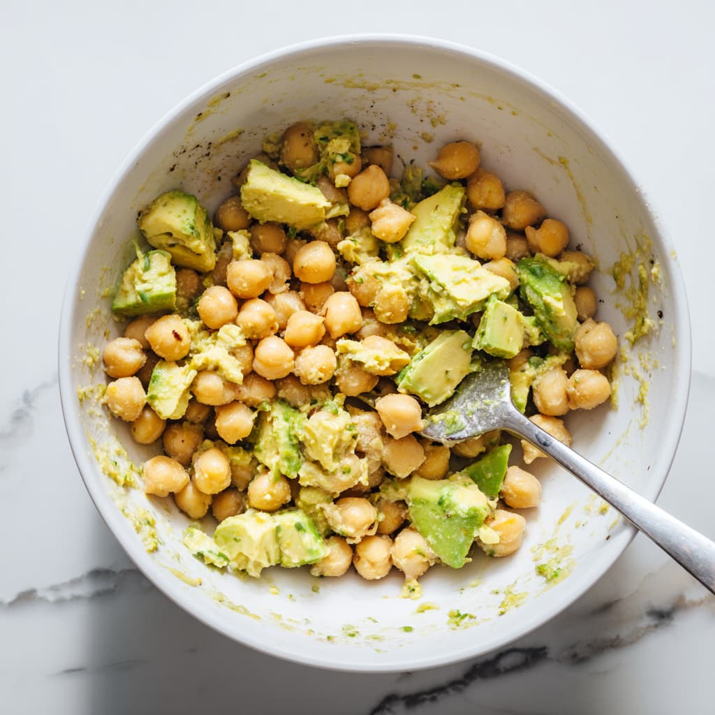 Chickpeas and ripe avocado being mashed together in a bowl with a fork to make creamy chickpea avocado salad filling.