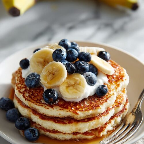 Stack of fluffy high-protein cottage cheese pancakes topped with fresh fruit, Greek yogurt, and a drizzle of maple syrup on a white plate.