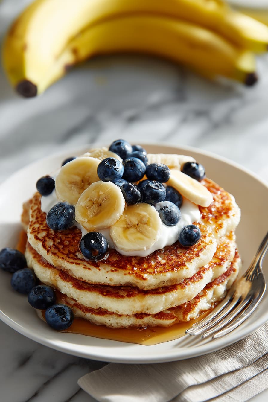 Stack of fluffy high-protein cottage cheese pancakes topped with fresh fruit, Greek yogurt, and a drizzle of maple syrup on a white plate.