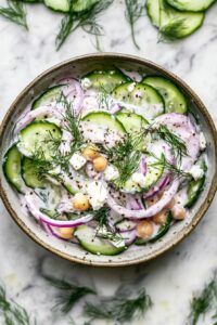 High protein cucumber salad with chickpeas, red onion, and Greek yogurt dressing, garnished with fresh dill and feta in a ceramic bowl