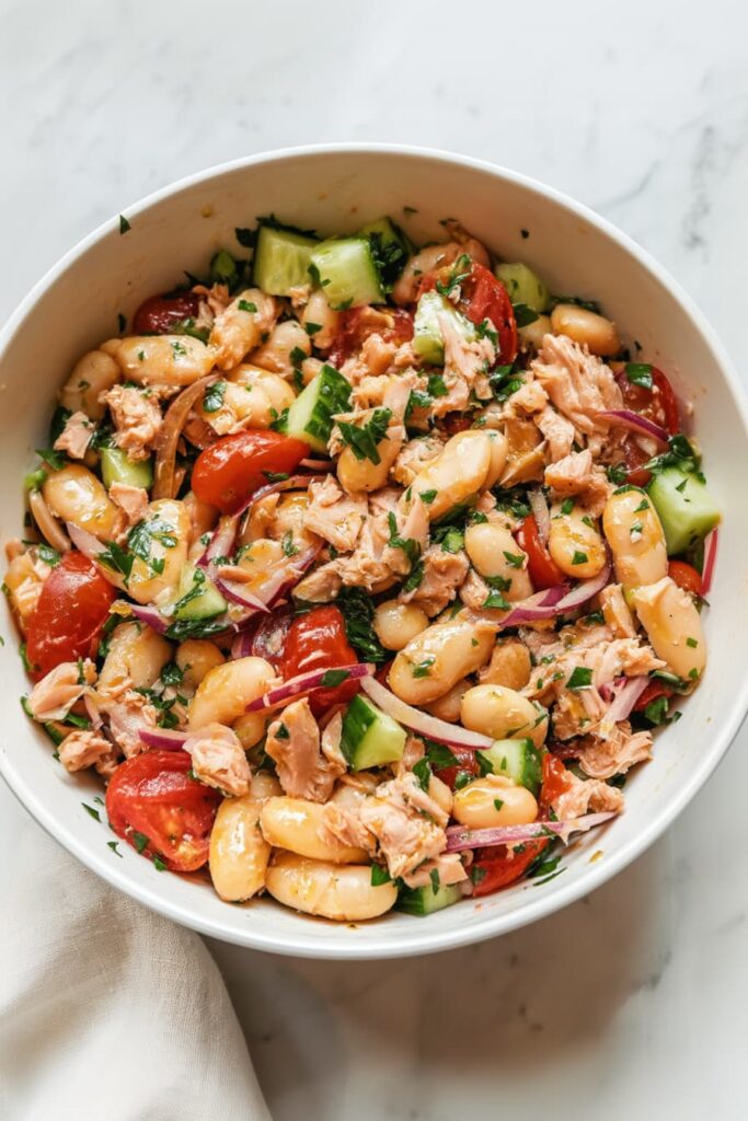 High protein tuna and white bean salad fully mixed in a white bowl with flaked tuna, cannellini beans, cherry tomatoes, cucumber, red onion, parsley, and lemon dressing, no mayo healthy meal