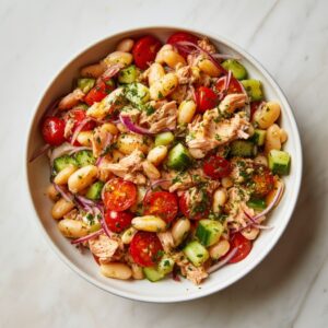 High protein tuna and white bean salad fully mixed in a white bowl with flaked tuna, cannellini beans, cherry tomatoes, cucumber, red onion, parsley, and lemon dressing, no mayo healthy meal