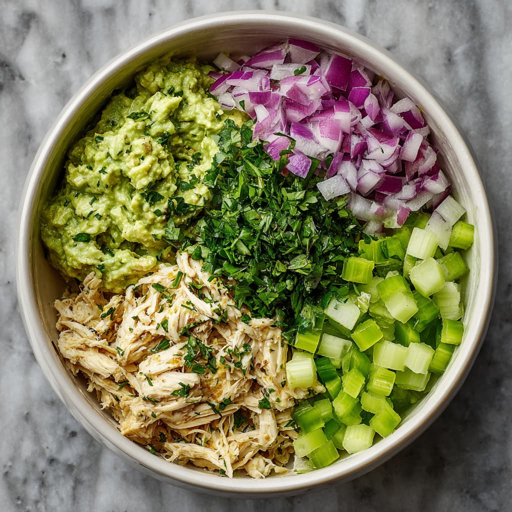 Shredded chicken, avocado dressing, celery, red onion, and herbs in a bowl, ready to be mixed for avocado chicken salad.