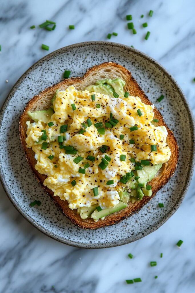 Scrambled eggs with cottage cheese and avocado toast on a white plate, garnished with chives and black pepper.