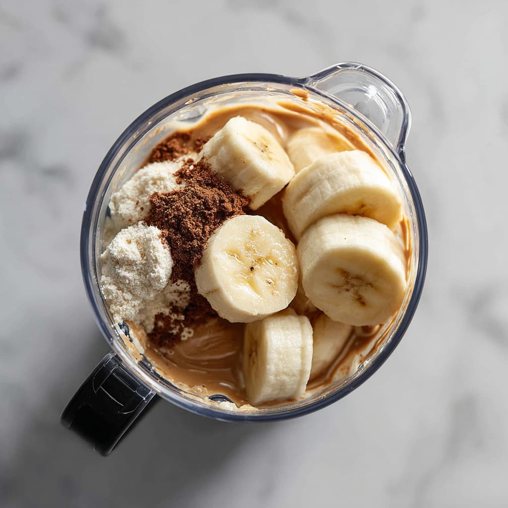 All smoothie ingredients including banana, protein powder, almond butter, and cold brew coffee added to a blender.