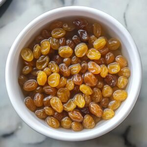 Golden raisins soaking in hot water in a small white bowl.
