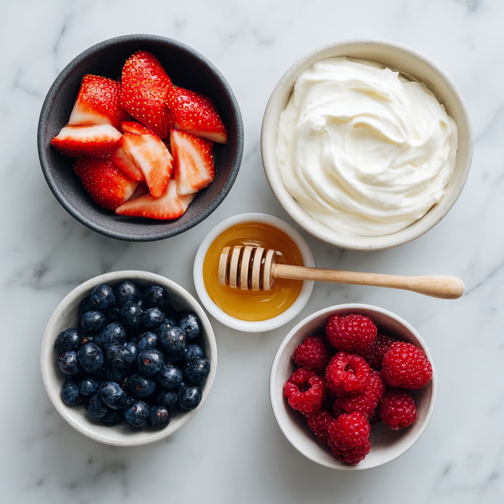 Bowl of Greek yogurt, sliced strawberries, blueberries, honey, and raspberries arranged on a white marble surface for making Greek yogurt bark.