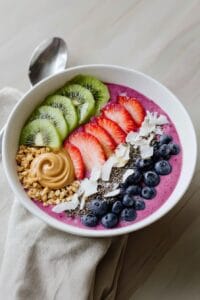 Tropical berry protein smoothie bowl topped with kiwi, berries, granola, chia seeds, and coconut, photographed overhead on a marble surface.