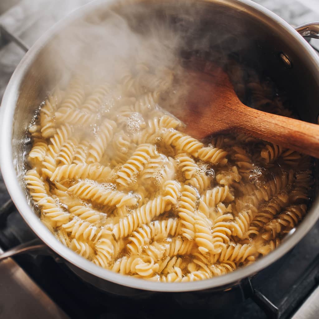 Whole wheat rotini pasta boiling in salted water in a stainless steel pot with steam rising and a wooden spoon on the edge.