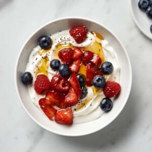 Greek yogurt bowl topped with sliced strawberries, blueberries, raspberries, honey drizzle and chia seeds on a marble surface