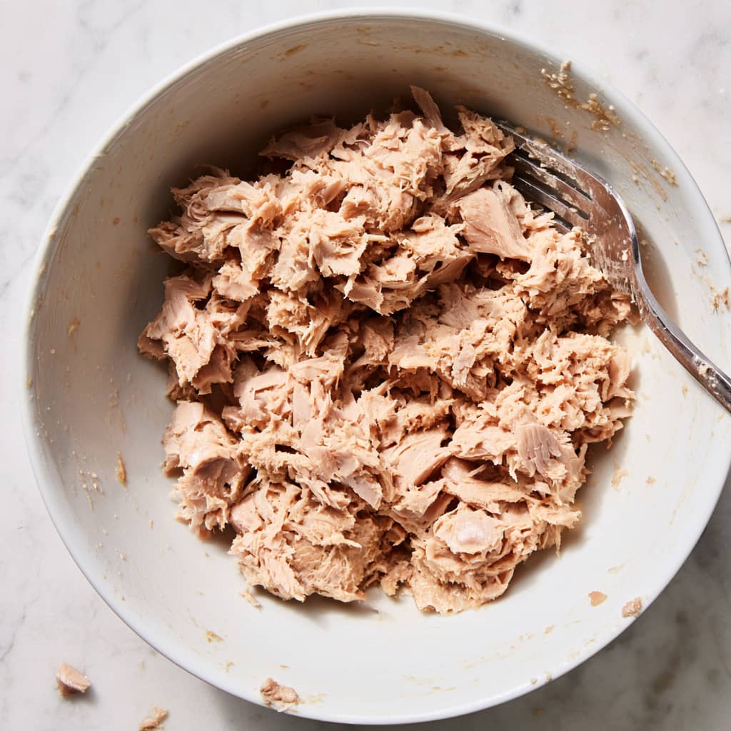 drained tuna flakes in a white bowl being broken apart with a fork to prepare tuna salad base
