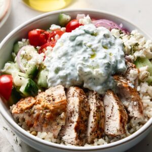 High protein Greek chicken bowl with grilled chicken, rice, fresh vegetables, feta cheese, and tzatziki sauce