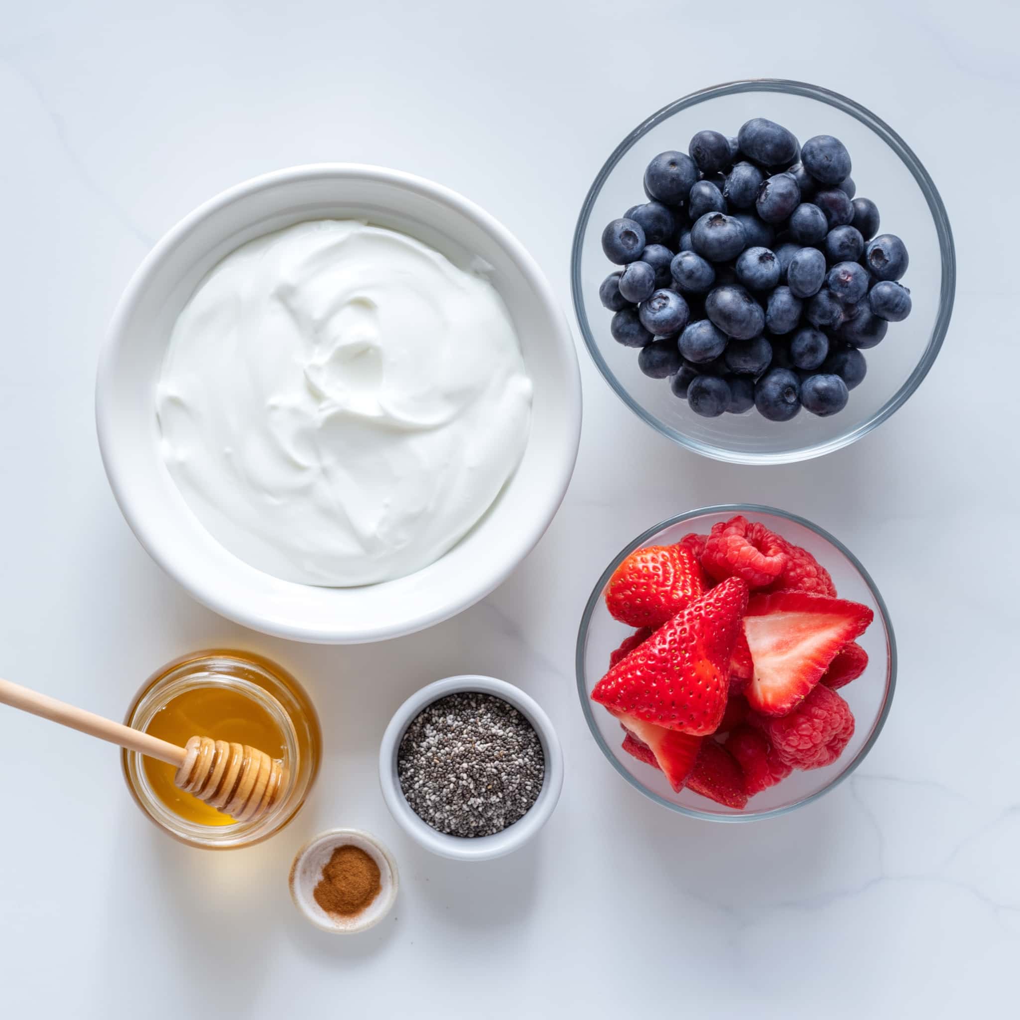 Ingredients for Greek yogurt with berries and honey including yogurt, blueberries, strawberries, raspberries, honey, chia seeds and cinnamon on a marble surface