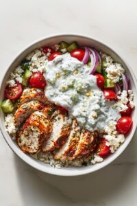 High protein Greek chicken bowl with grilled chicken, rice, fresh vegetables, feta cheese, and tzatziki sauce