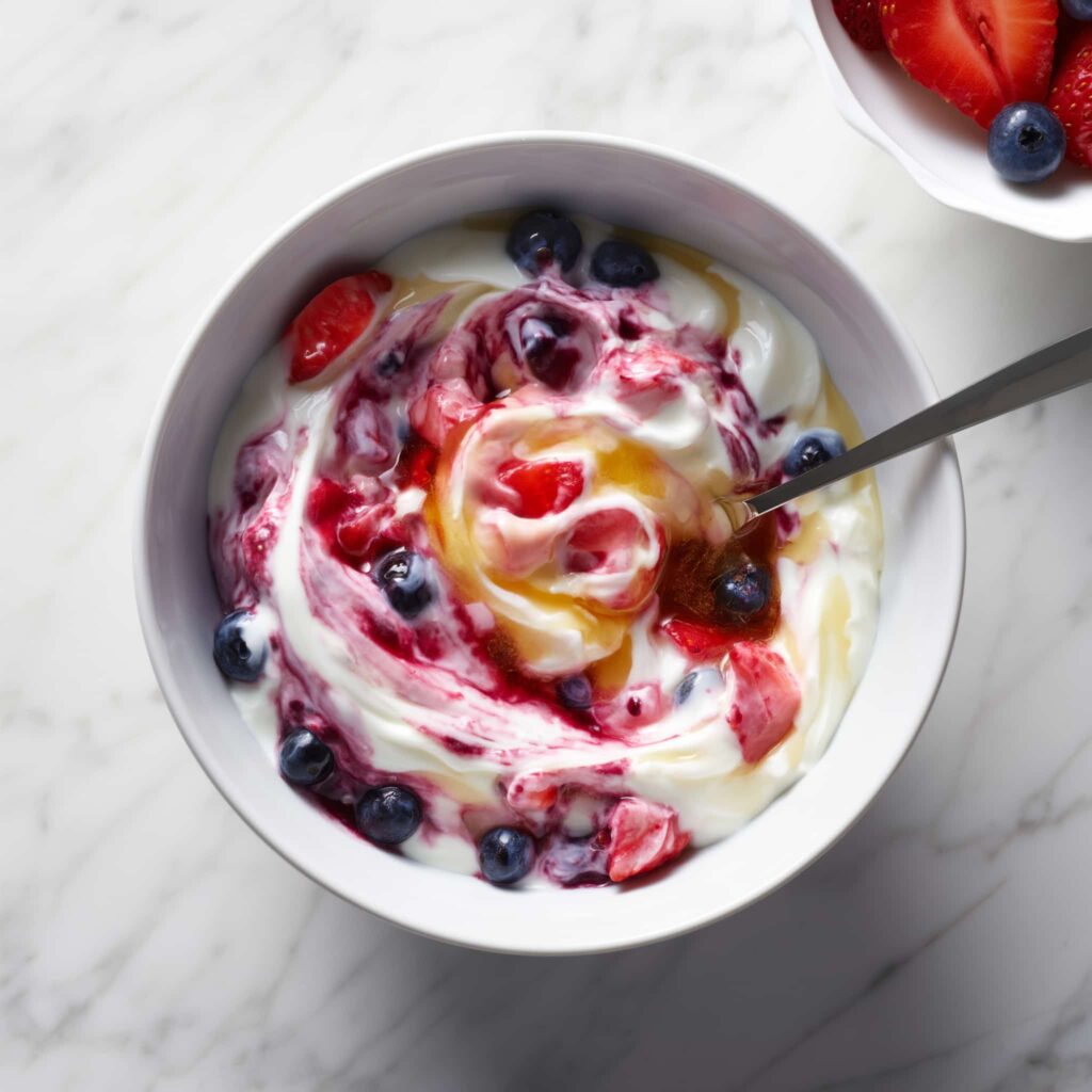 Greek yogurt mixed with berries and honey showing swirled texture with strawberries and blueberries in a white bowl