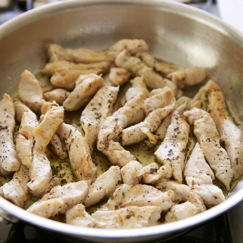Sliced chicken breast strips cooking in a stainless steel pan with light seasoning and slight browning