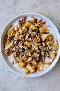 Healthy cinnamon apple yogurt bowl topped with warm apples, granola, walnuts, oats, and chia seeds in a white bowl.