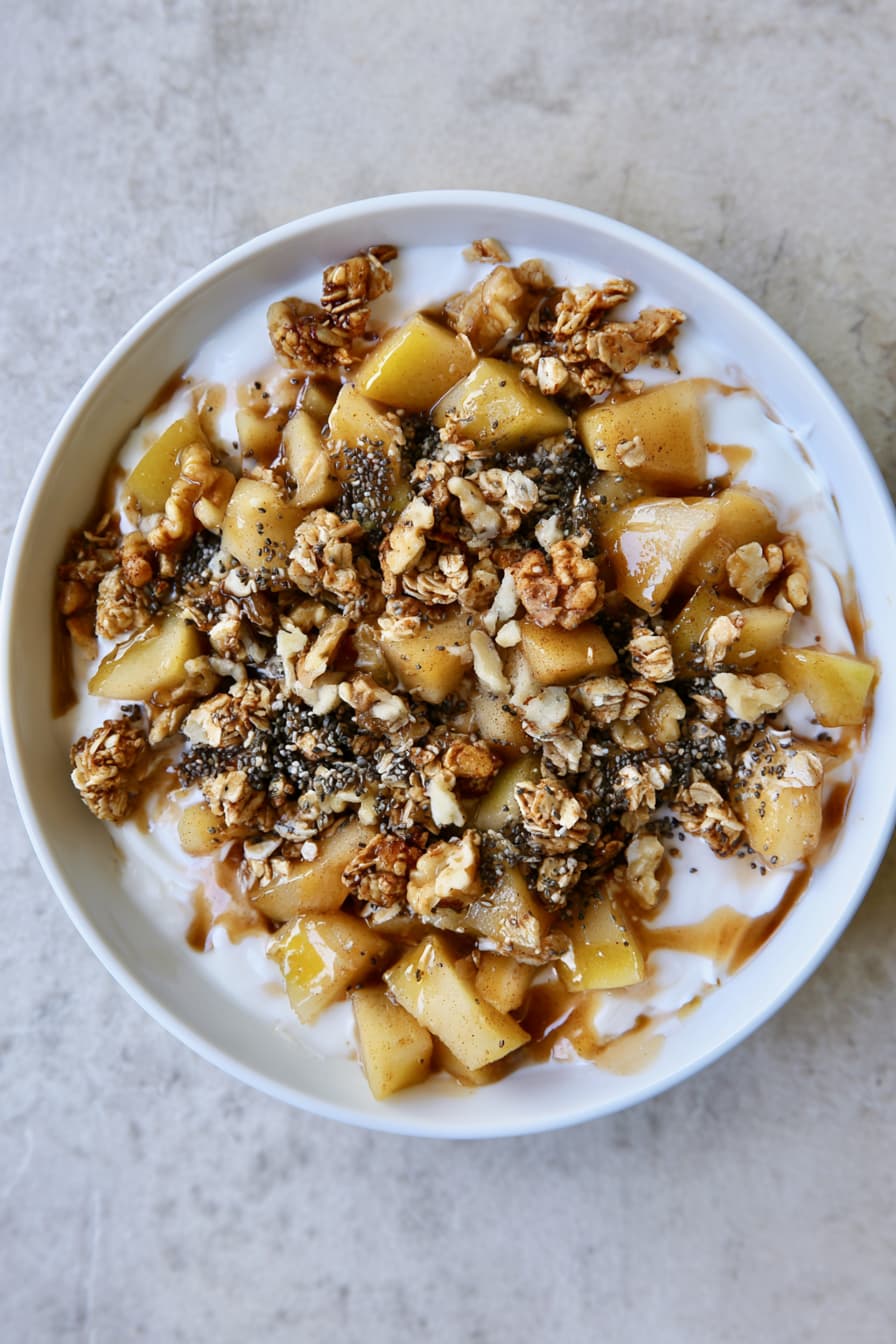 Healthy cinnamon apple yogurt bowl topped with warm apples, granola, walnuts, oats, and chia seeds in a white bowl.