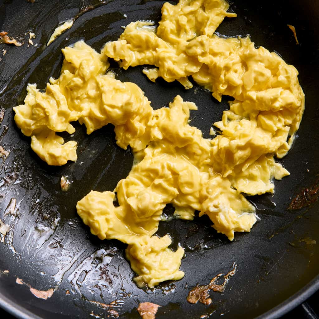 Soft scrambled egg curds cooking in a dark skillet with browned bits from chicken breast pieces.