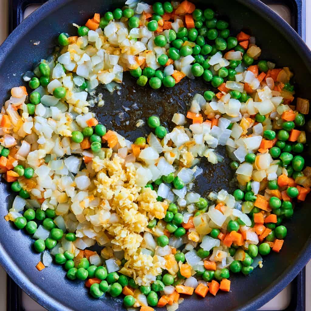 Diced onions, garlic, ginger, peas, and carrots sautéing in a skillet on the stovetop.