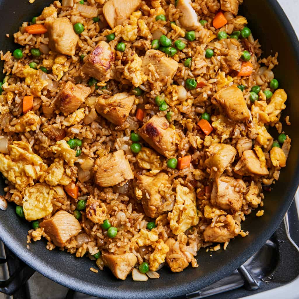 Chicken breast pieces, scrambled eggs, vegetables, and rice mixed with sauce in a skillet.