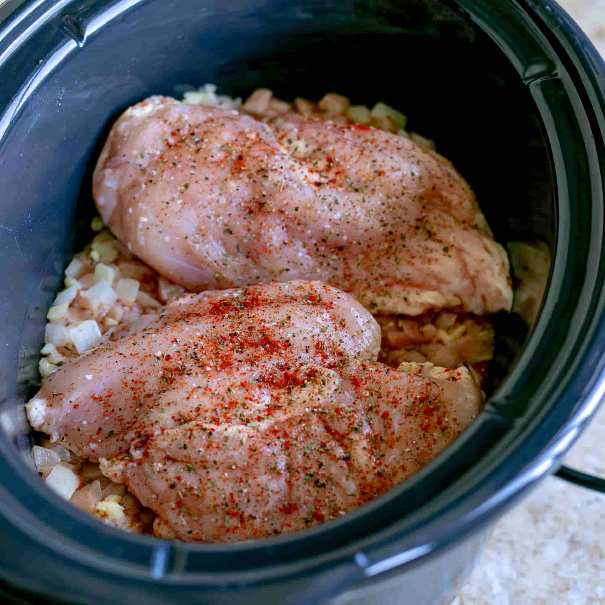 Seasoned raw chicken breasts placed into the crockpot over onions and garlic.